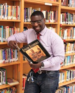 Augustine Obeng standing by a book shelf holding an open book entitled African Beginnings