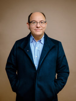 Portrait of Sakis Gekas, standing, hands in coat pockets, smiling