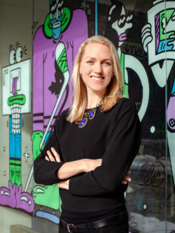 professor Lyndsay Hayhurst, smiling, arms folded, in front of wall of graffiti art