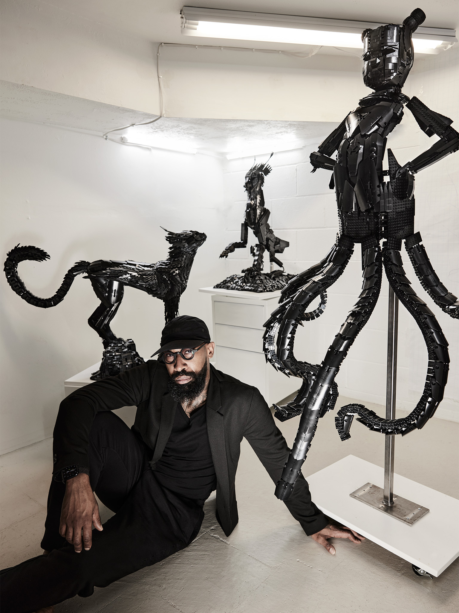 Ekow Nimako seated on the floor of his studio, three sculptures behind him