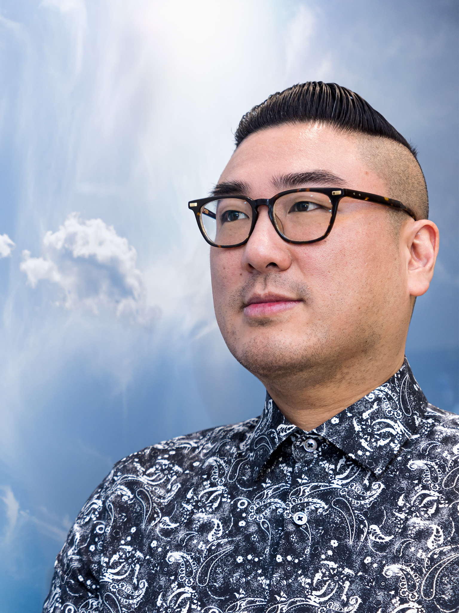 Michael Mak in paisley shirt, looking to the left, in front of blue sky with clouds background