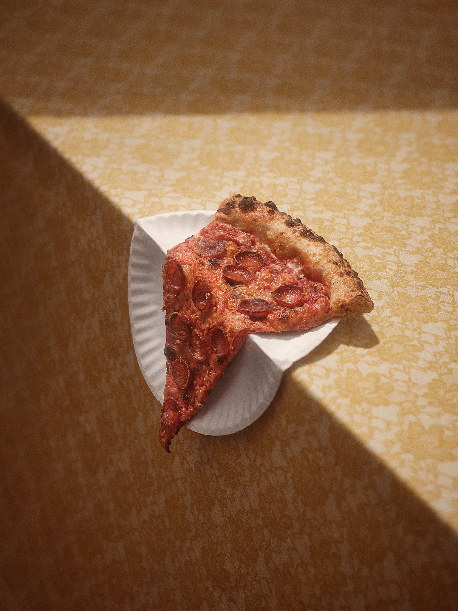 photograph of pizza slice on paper plate drooping off of the edge of a table