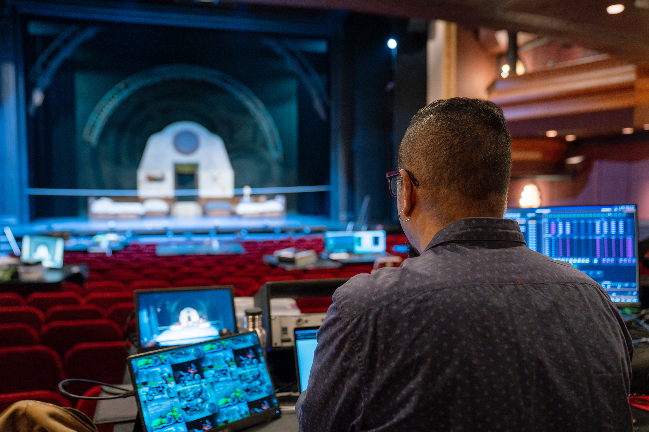 Arun Srinivasan seen from behind seated at lighting control table with theatre stage in the background